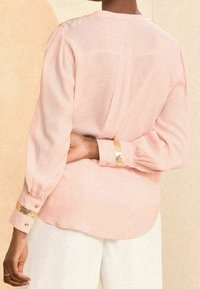 Blusa rosa chiaro con maniche lunghe, caratterizzata da dettagli arricciati sulle spalle e polsini con accenti dorati. Tessuto liscio con chiusura sul retro a bottoni.