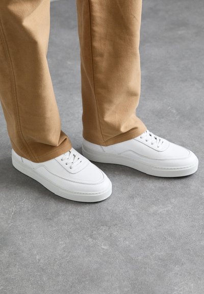 Sneakers bianchi in pelle con accenti testurizzati, suole in gomma piatta e lacci rotondi. Indossati con pantaloni beige, su uno sfondo grigio.