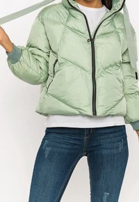 Lys grøn quilted jakke med hætte, frontlynlås, ribbede manchetter og snøre. Båret over en hvid skjorte og blå jeans.