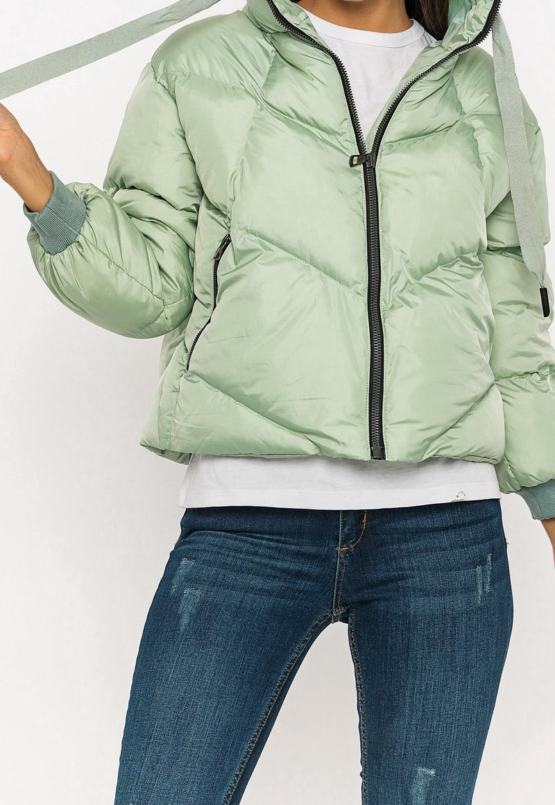 Lys grøn quilted jakke med hætte, frontlynlås, ribbede manchetter og snøre. Båret over en hvid skjorte og blå jeans.
