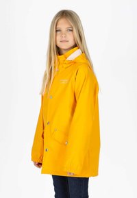 Jeune fille aux longs cheveux blonds portant une veste de pluie jaune vif à capuche et un jean bleu marine, debout devant un fond uni.