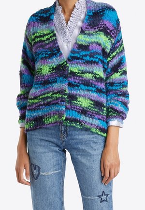 Personne portant un cardigan en maille épaisse à rayures bleues, vertes, violettes et noires, par-dessus une chemise à col à volants et un jean orné de patchs en forme d'étoiles et de cœurs.