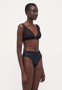 Bralette in pizzo nero con scollatura profonda e spalline larghe, abbinata a un paio di bikini neri a vita alta con una texture liscia.