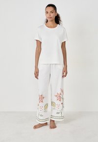 Weißes Baumwoll-T-Shirt mit hochgekrempelten Ärmeln, kombiniert mit weit geschnittenen Leinenhosen, die am Saum und in den Taschen mit bunten Blumenstickereien verziert sind.