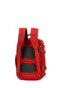 Mochila de tela roja con respaldo de malla acolchada, correas ajustables y lazos laterales para accesorios. Incluye un bolsillo con cremallera y hardware minimalista.