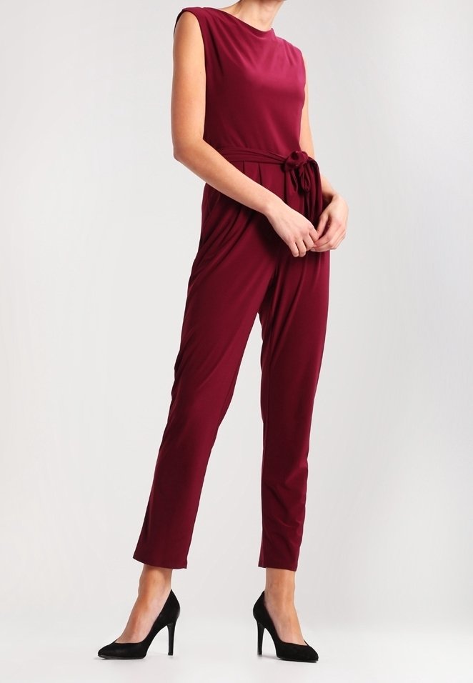 Combinaison bordeaux sans manches avec ceinture nouée à la taille, pantalon droit, portée avec des chaussures à talons noirs sur fond uni.