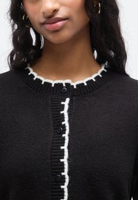 Μαύρο πλεκτό ζακέτα με λευκή γιακαδάκι σε σχήμα κυματιστού, με κούμπωμα με κουμπιά μπροστά και υφασμάτινη υφή. Απλός σχεδιασμός με αντιθετικές λεπτομέρειες.