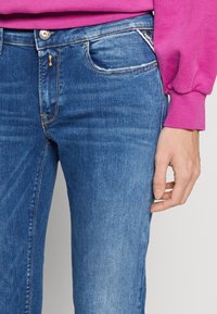 Vaqueros de mezclilla azul de corte slim, con cinco bolsillos, costuras en contraste y una pequeña etiqueta en el bolsillo delantero. Textura suave.