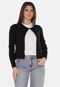 Cardigan nero con texture a costine, scollatura rotonda e design aperto. Abbinato a una camicia bianca e jeans azzurri chiari.