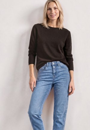 Femme aux cheveux blonds portant un haut noir à manches longues et un jean taille haute bleu clair, debout contre un mur clair uni.