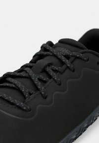 Närbild på svart sneaker med mönstrade svarta skosnören och vågig design längs sidan, visar vadderad plös och del av sulan.