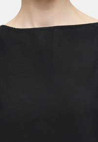 Κοντινό πλάνο ενός ατόμου που φοράει μαύρο τοπ με λαιμόκοψη boat neck, εμφανίζοντας το λαιμό και το άνω μέρος του στήθους πάνω σε απλό φόντο.