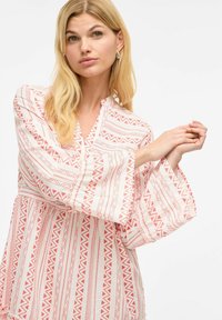 Blouse rouge et blanche à motif, avec un décolleté en V, des manches longues fluides et un tissu texturé présentant des motifs géométriques.