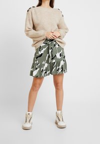 Vrouw in een beige gebreide trui, met een groen-witte camouflage rok met ruffles en witte hoge sneakers, staande tegen een effen achtergrond.