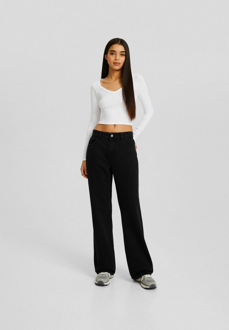 Jeans neri a gamba larga in denim, abbinati a un top aderente a maniche lunghe bianco. L'outfit è completato da sneakers grigie.