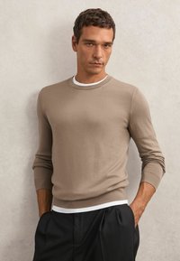 Jasnobrązowy sweter dzianinowy z okrągłym dekoltem i długimi rękawami, noszony na białej koszuli. Materiał wydaje się gładki z ściągaczami na mankietach.