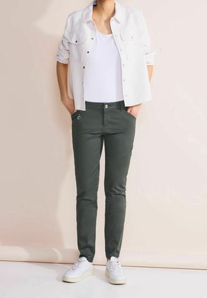 Personne portant un pantalon slim vert foncé, des baskets blanches, un débardeur blanc et une chemise blanche à manches retroussées, les mains dans les poches.