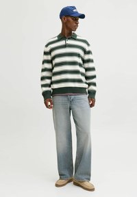 Svītrainā zaļā un baltā pulovera džemperis, brīvi pieguļoši gaiši zili džinsi, bēša apavi un zils cepure ar logotipa detaļu.