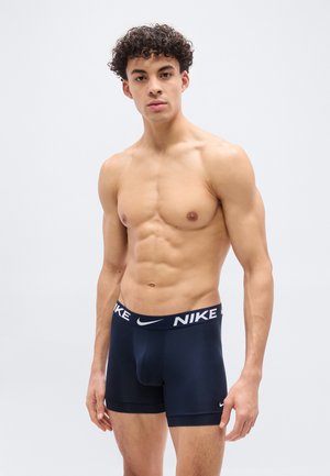 Ευχάριστος νέος άνδρας με σγουρά μαλλιά, φορώντας σκούρα μπλε μποξέρ της Nike, στέκεται μπροστά σε απλό ανοιχτό φόντο.