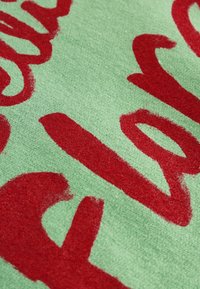 Grønt stof med en blød tekstur, der har store, røde håndmalede bogstaver. Designet inkluderer buede og flydende tekstmønstre.