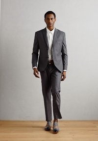 Completo grigio su misura con vestibilità slim, camicia bianca e cintura marrone. Il modello indossa scarpe grigie chiare con punta rotonda, in piedi su pavimento in legno.
