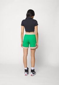 Zkrácená černá polokošile s krátkými rukávy, spárovaná se zelenými a béžovými sportovními šortkami s bočními detaily, nošená s černými vysokými teniskami.