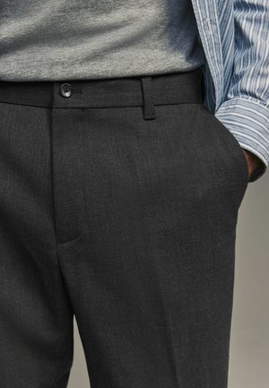 Pantalones gris oscuro hechos de tela texturizada, con cierre de botón, trabillas para cinturón y bolsillos laterales. Combinados con una camisa gris y una chaqueta a rayas.