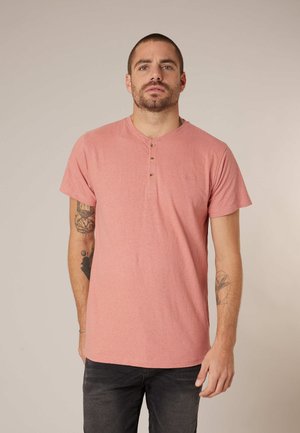 Mann mit kurzen Haaren und Tätowierungen, der ein lachsfarbenes Henley-Shirt mit kurzen Ärmeln und dunkle Hosen trägt und vor einem schlichten Hintergrund steht.