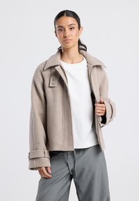 Woman with dark hair tied back wearing beige zip-up jacket over white shirt and loose gray pants, standing against plain light background.