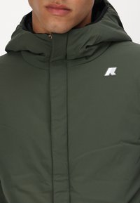 Veste à capuche verte en tissu lisse. Équipée d'une fermeture éclair et d'un accent logo blanc sur la zone de la poitrine gauche.