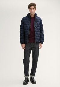 Marineblaue Steppjacke mit hohem Kragen, Reißverschluss und Seitentasche, getragen über einem burgunderfarbenen Pullover und dunklen Jeans mit aufgerollten Säumen.