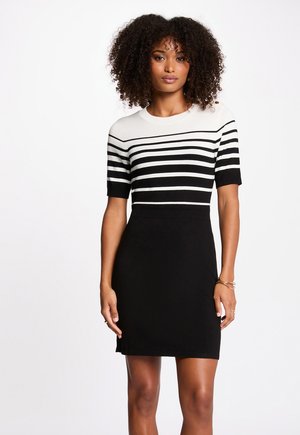 Kvinde med krøllet hår iført en sort-hvidstribet kortærmet kjole, der står foran en ensfarvet hvid baggrund.