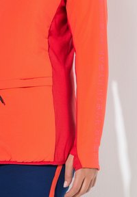 Jasnoczerwona kurtka z kontrastującymi ciemnoczerwonymi akcentami. Posiada kieszeń na zamek i nadrukowany tekst wzdłuż rękawa. Gładka tekstura.