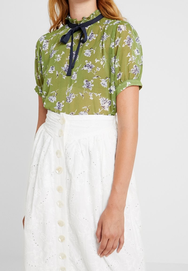 Femme portant un chemisier à manches courtes vert à fleurs avec un nœud papillon noir et une jupe blanche taille haute boutonnée avec motif œillet.