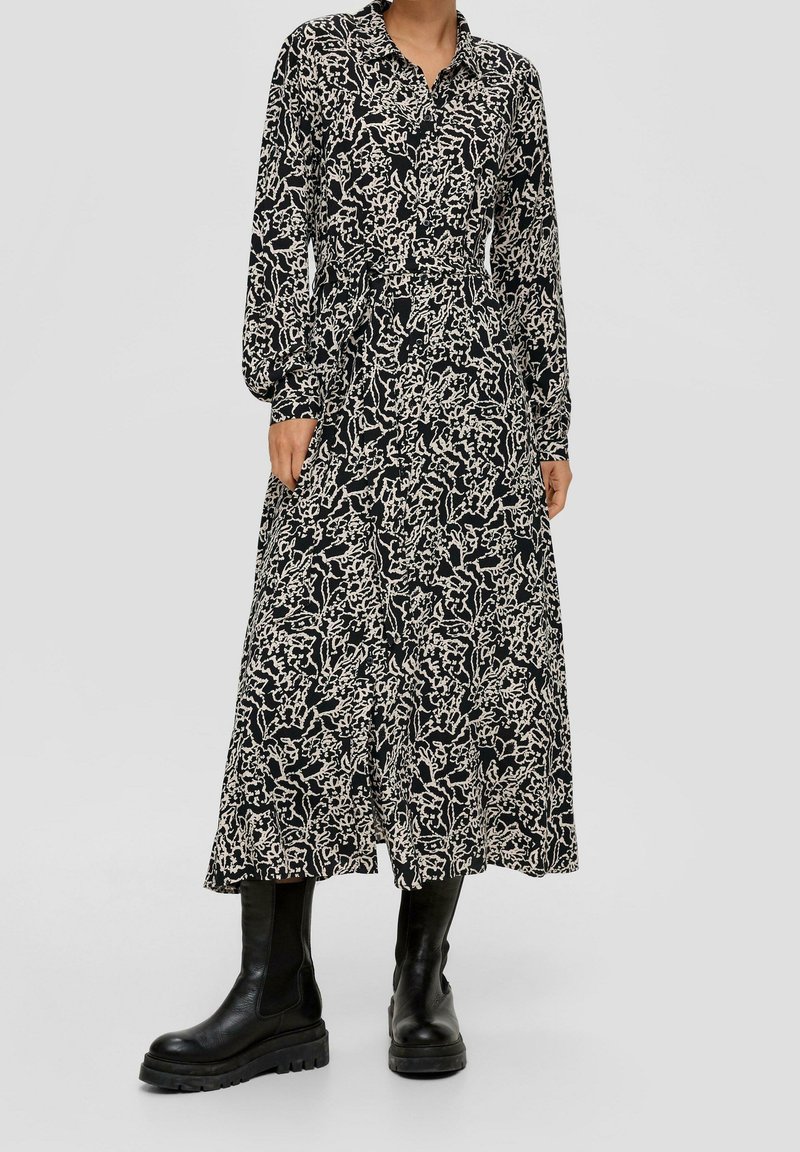 Femme portant une robe midi boutonnée à manches longues avec un motif floral noir et blanc, associée à des bottes en cuir noires épaisses.
