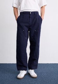 Mørkeblå, løstsiddende bukser med sidelommer, knaplukning og sølvfarvede hardware-detaljer. Båret med hvide sneakers på en struktureret gulv.