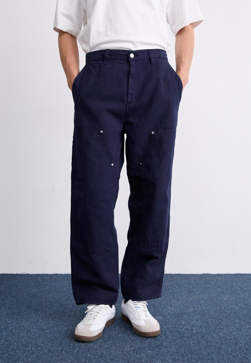 Marineblaue, locker sitzende Hose mit Seitentaschen, Knopfverschluss und silbernen Hardware-Akzenten. Getragen mit weißen Sneakers auf einem strukturierten Boden.