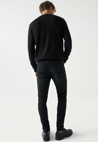 Suéter de punto negro y jeans negros ajustados, vistos desde atrás. Botas con suela texturizada. Tejido liso y líneas limpias en el diseño.