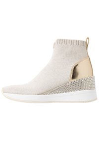 Beige pletená kotníková bota s kovově zlatým podpatkem a dekorativními cvočky. Má texturovaný svršek a bílou gumovou podrážku.