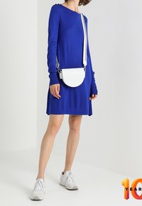 Frau in einem blauen Langarmkleid, mit einer weißen Umhängetasche und weißen Sneakers, steht vor einer weißen Wand.