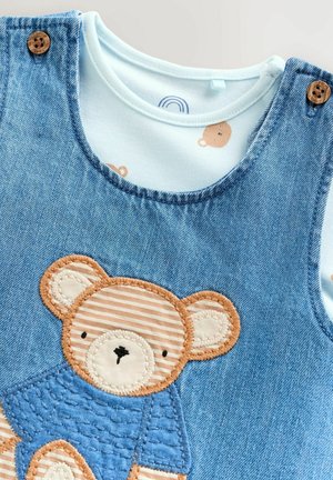 Džínové kombinézy s modrou nášivkou medveďa. Svetlo modrá vnútorná košeľa má vzor malých medveďov. Drevené gombíky zvýrazňujú popruhy.