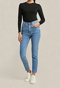 Femme portant un haut noir à manches longues, un jean skinny taille haute bleu clair et des baskets blanches, debout avec une main dans la poche.