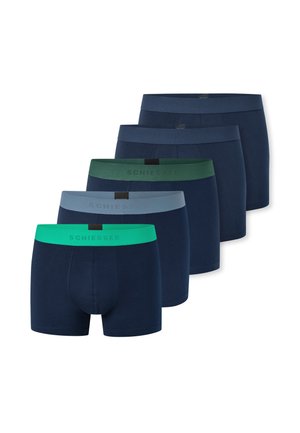 Skjorteløs mand med mørkt hår iført marineblå boxershorts med grøn taljebånd, poserer med den ene hånd på nakken og den anden på hoften.