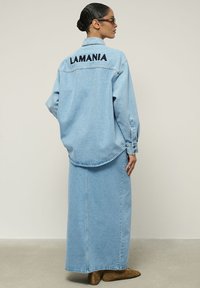 Šviesiai mėlyna džinsinė marškinė su žodžiu "LAMANIA" išsiuvinėtu ant nugaros, derinami su atitinkančiomis ilgu sijonu ir rudos spalvos auliniais batais iki kulkšnies.