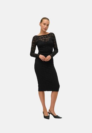 Svart spetsklänning med långa ärmar, tajt bodice och knälång kjol. Kombinerad med svarta slingback-klackar med rosettdetaljer.