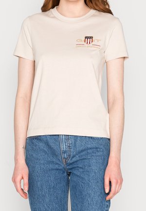 Beżowy t-shirt z krótkim rękawem z logo "GANT 1949" i emblematem w kształcie tarczy na piersi, noszony z niebieskimi dżinsami przez stojącą osobę.