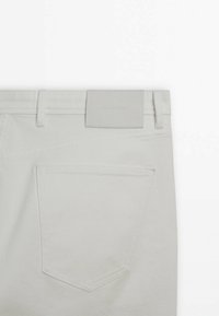 Vue de dos d'un pantalon en denim beige clair montrant la poche arrière, les passants de ceinture et une étiquette de marque rectangulaire cousue à la taille.