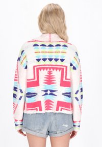 Färgglad stickad tröja med geometriska mönster i rosa, blått och grönt, med en lös passform och mjuk textur, bärs över denimshorts.