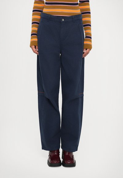 Personne portant un pantalon large bleu marine, un haut à rayures marron et orange à manches longues, et des chaussures brillantes rouge foncé, se tenant devant un fond blanc.