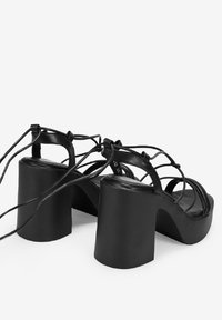 Sandalias negras de tacón alto con un chunky block heel, diseño de tiras y cierre ajustable con cordones. Textura de cuero suave y estilo minimalista.